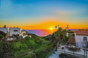 Vista costeira com pôr do sol, um caminho que leva a um edifício com área de estar ao ar livre e zona de piscina.
