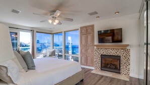A bedroom with a large bed, a fireplace, a ceiling fan, and a TV.
