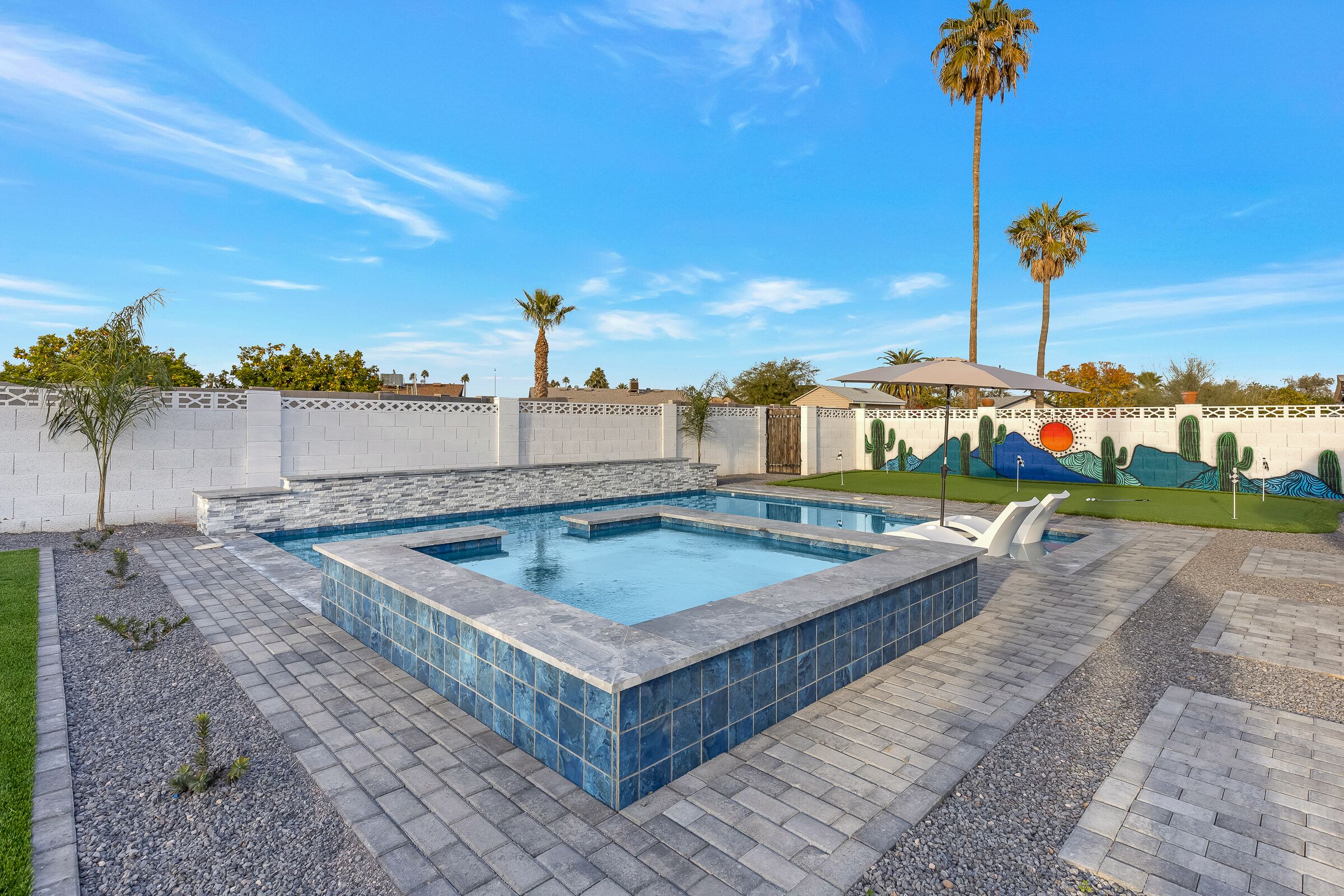 A rectangular swimming pool with a stone border, surrounded by a paved patio area with a low wall and palm trees.