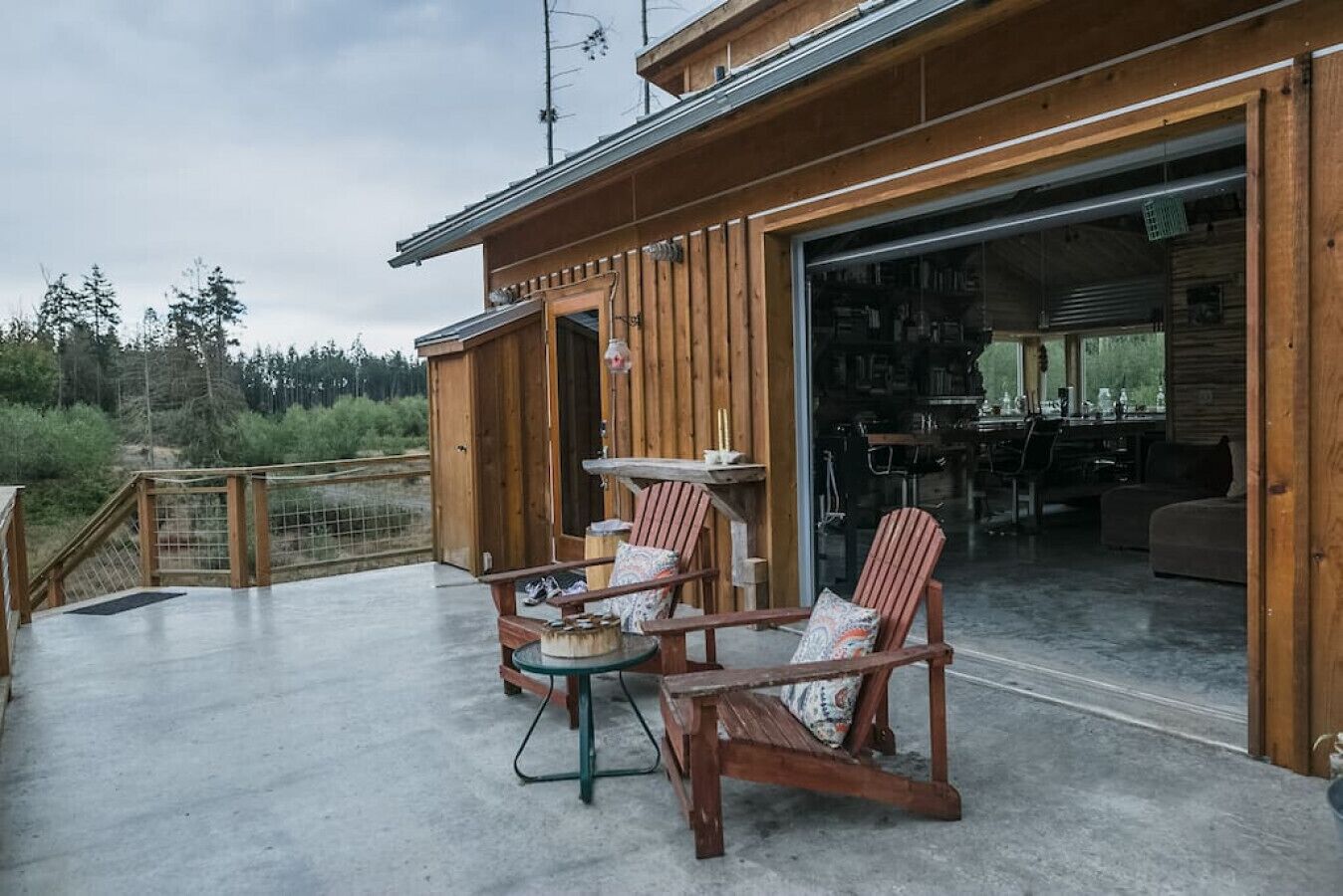 A wooden cabin with a patio area featuring a table and chairs, a large sliding door leading to an interior space, and a view of the surrounding forest.
