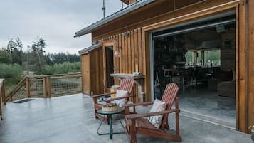 A wooden cabin with a patio area featuring a table and chairs, a large sliding door leading to an interior space, and a view of the surrounding forest.