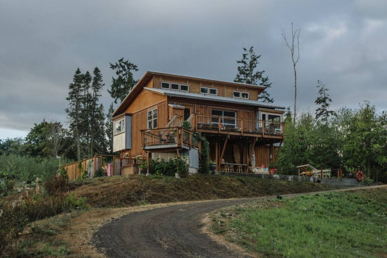 Une maison en bois à deux étages, dotée d’une terrasse, entourée d’arbres et bordée d’un sentier de terre.