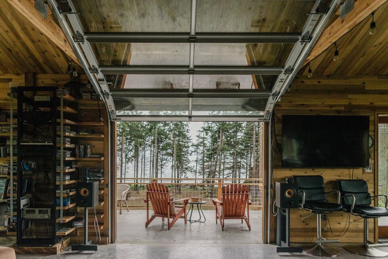 L’intérieur d’un chalet accueillant, avec une grande fenêtre donnant sur une forêt, des murs en bois, une bibliothèque et un téléviseur.