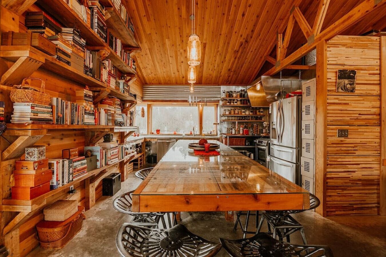 A wooden interior with a central island table, bookshelves, and a kitchen area.