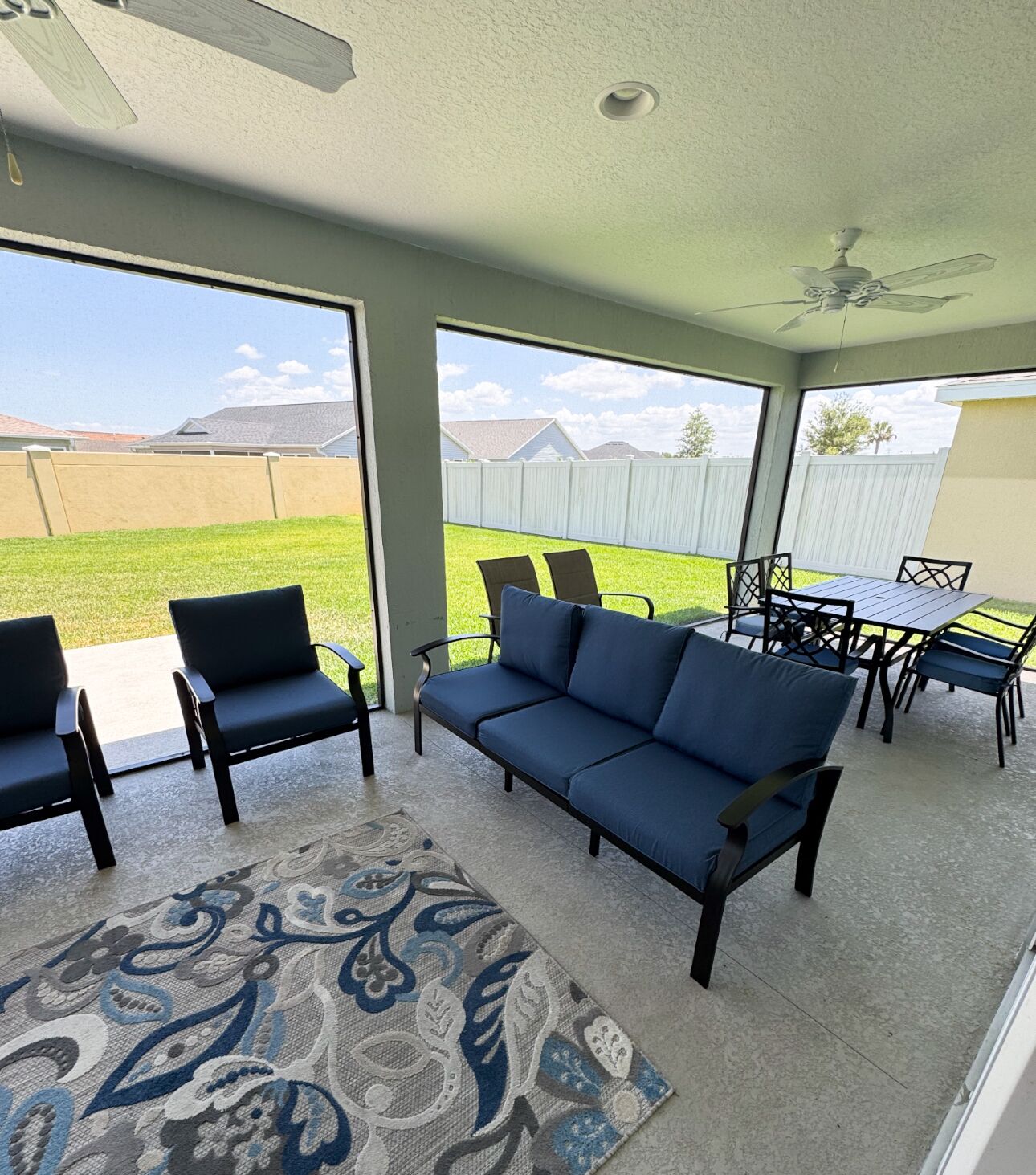 Ein überdachter Patio-Bereich mit einem blauen Sofa, zwei Stühlen, einem Esstisch und einem Deckenventilator.