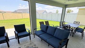 Ein überdachter Patio-Bereich mit einem blauen Sofa, zwei Stühlen, einem Esstisch und einem Deckenventilator.