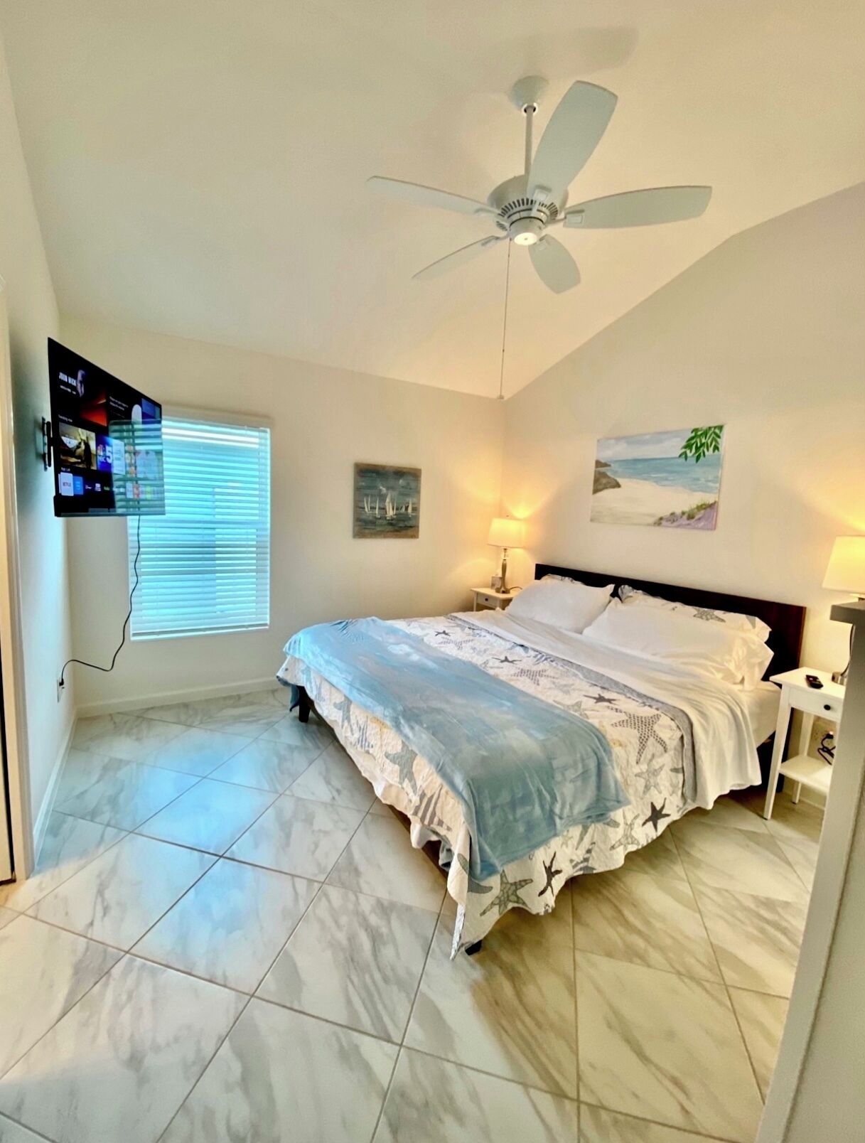 A bedroom with a bed, a ceiling fan, a TV mounted on the wall, and artwork on the walls.