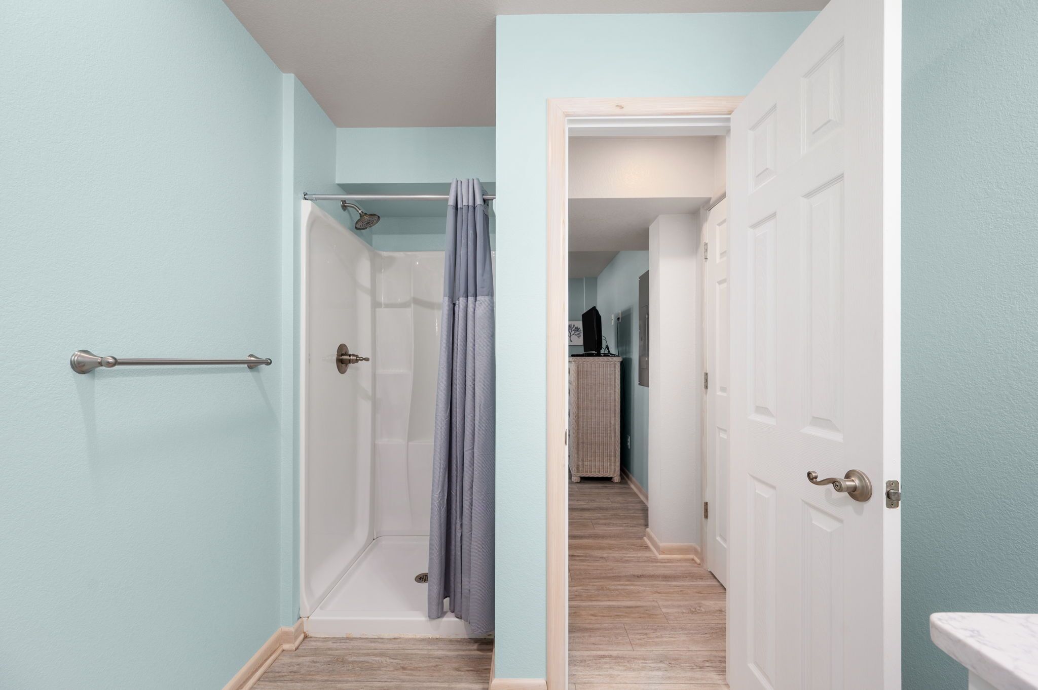 A narrow hallway with a shower area, a door leading to a room with a sink, and a wooden floor.