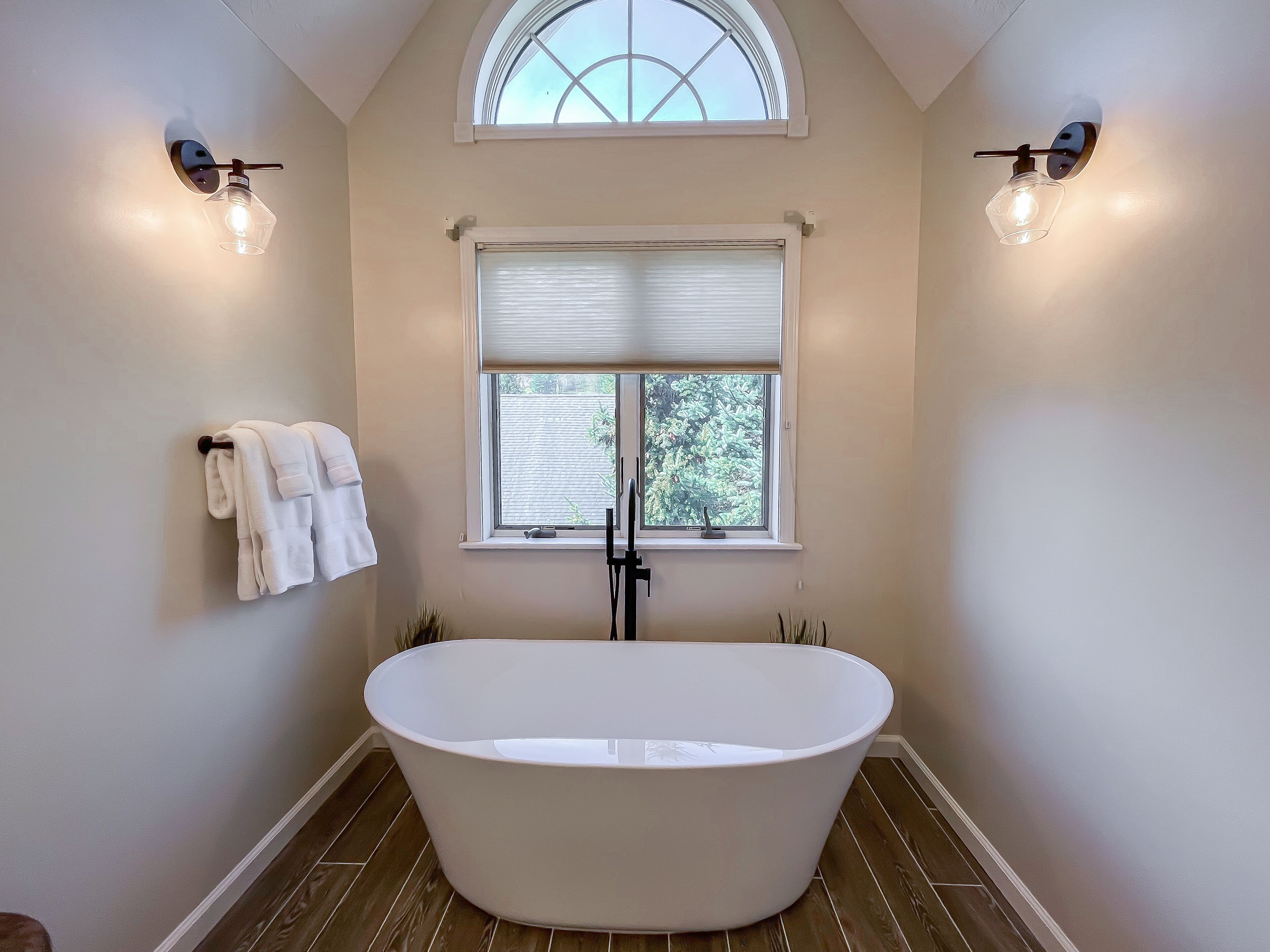 A bathroom with a freestanding bathtub, a window with blinds, and two wall-mounted light fixtures.