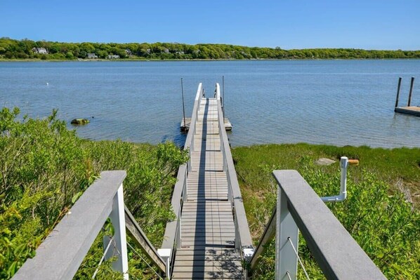 A wooden dock leading into a calm lake with a grassy area on one side and a row of houses in the background. - Waterfront with Private Dock! (Eastham)