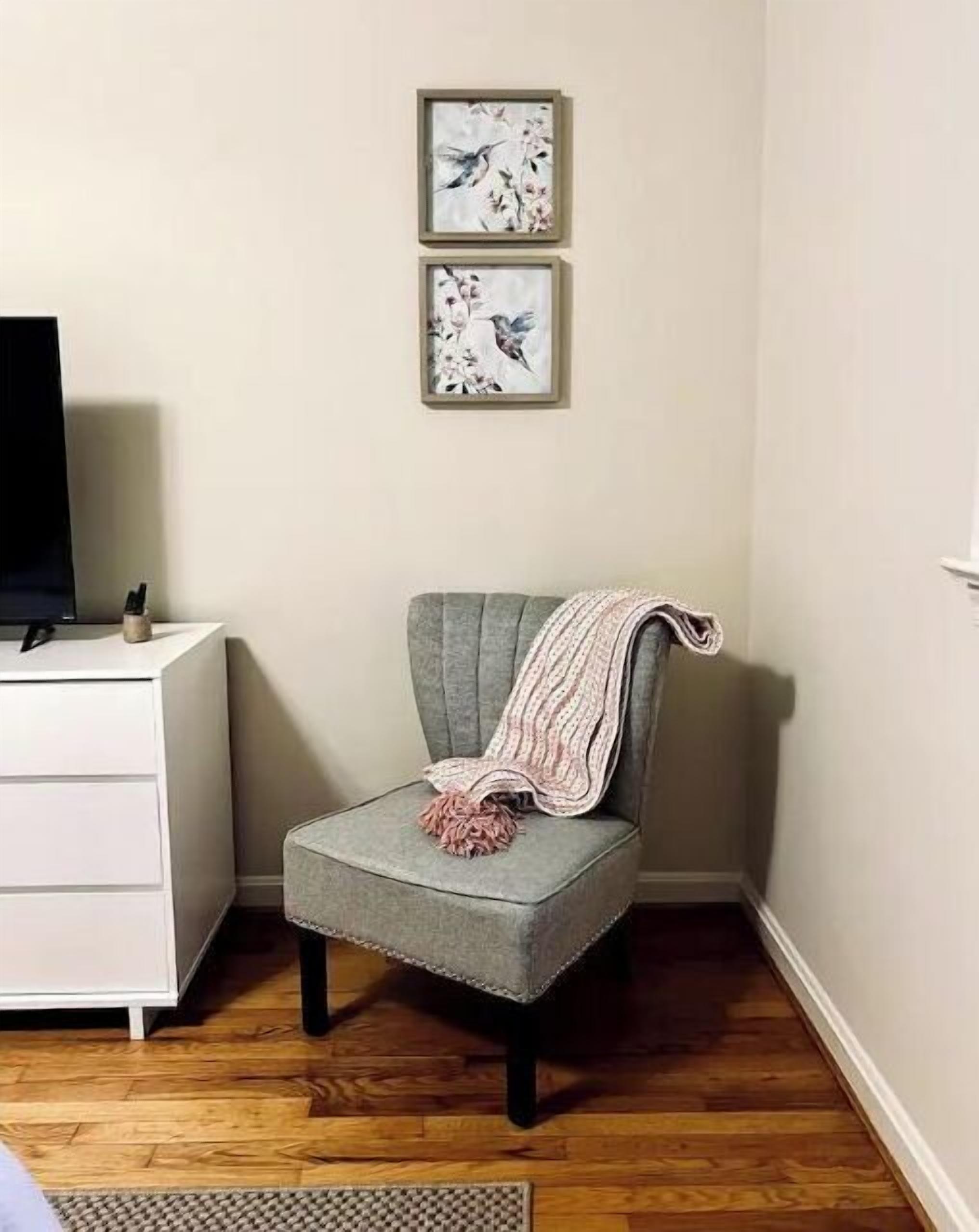 A room with a grey armchair, a white dresser, and two framed pictures on the wall.