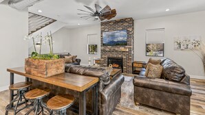 A modern living room with a brick fireplace, leather sofas, a wooden dining table with stools, and a ceiling fan. - "The Spruce"-Gathering Tree is your Ultimate Family Reunion Spot (Flagstaff)