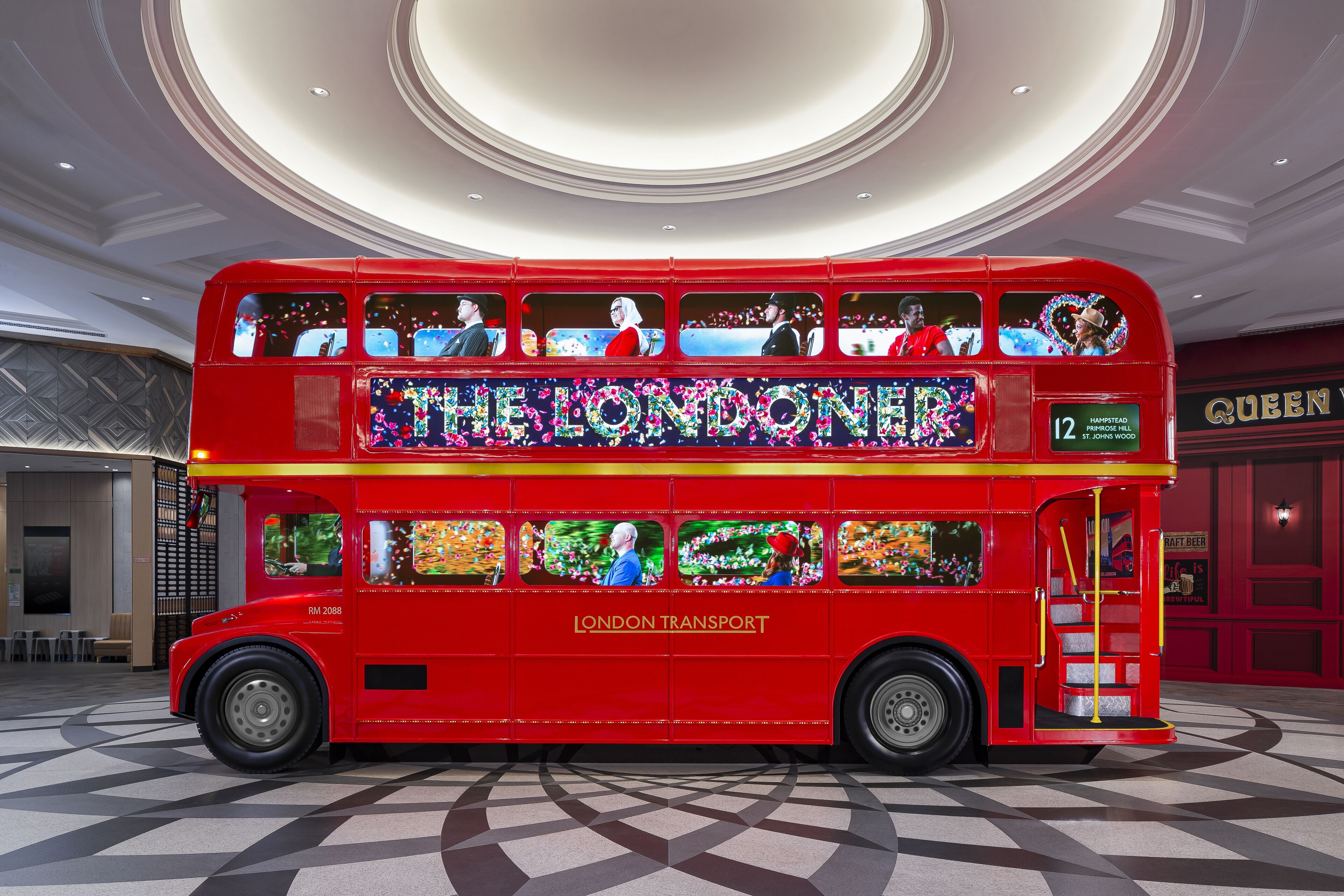 A red double-decker bus with "The Londoner" and "London Transport" on the side, parked indoors with a checkered floor and a circular ceiling design.