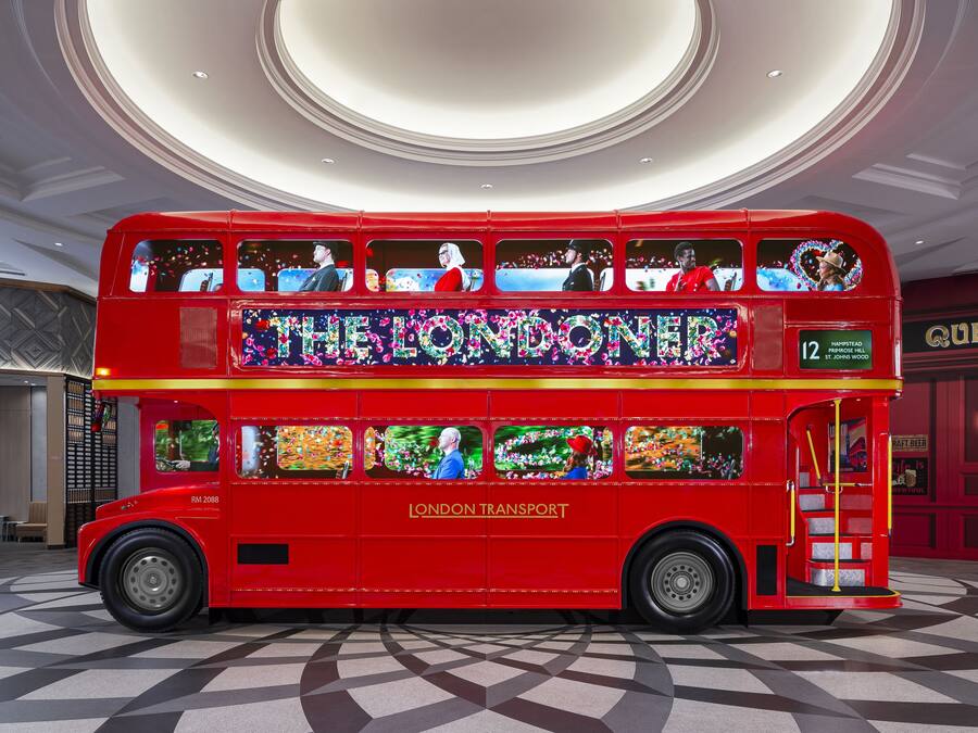A red double-decker bus with "The Londoner" and "London Transport" on the side, parked indoors with a checkered floor and a circular ceiling design.