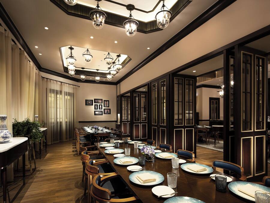 A spacious dining area with a long table set for a meal, elegant chandeliers, and a bar area visible through an open doorway.