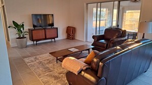 A living room with a leather sofa, a coffee table, a TV on a cabinet, and a large window.