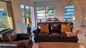 A living room with a brown leather sofa, a matching armchair, and a wooden coffee table. There are two floor lamps and a large window providing a view of the outdoors.