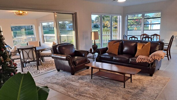 A spacious living room with a leather sofa, armchairs, and a coffee table. There is a dining area with a table and chairs, and large windows offering a view of the outdoors.