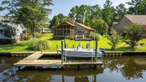 A house with a dock extending into a calm body of water, surrounded by greenery and trees. - Waterfront Retreat (New Bern)