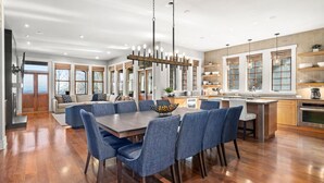 Un vaste espace de salle à manger avec une grande table en bois et des chaises recouvertes d’un tissu bleu, une cuisine moderne dotée de meubles en bois et d’appareils électroménagers en acier inoxydable, et de grandes fenêtres offrant une vue sur l’extérieur.