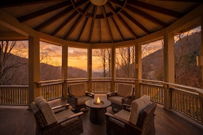 A covered outdoor seating area with wicker chairs and a central fire feature, overlooking a mountainous landscape. - Whisper Mountain Lodge | Luxury Mountain Rental with Hot Tub, Fire Pit, Game Room + Views (Leicester)