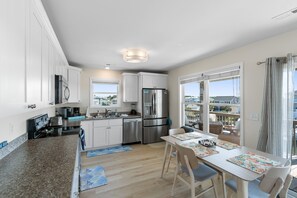 A modern kitchen with white cabinets, stainless steel appliances, and a dining area with a table and chairs. - NEW to PROACTIVE Vacations! "Tri-D's" is a stunning 6 bedroom, 4 bathroom home WITH elevator! (Holden Beach)