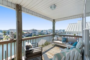 A covered deck with a wooden railing, a cushioned seating area, and a view of the marina and houses in the background. - NEW to PROACTIVE Vacations! "Tri-D's" is a stunning 6 bedroom, 4 bathroom home WITH elevator! (Holden Beach)