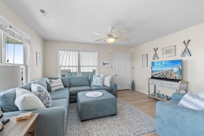 A living room with a sectional sofa, ottoman, flat-screen TV, ceiling fan, and a wall-mounted TV stand. - NEW to PROACTIVE Vacations! "Tri-D's" is a stunning 6 bedroom, 4 bathroom home WITH elevator! (Holden Beach)