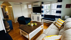 A cozy living room with a sofa, a coffee table, and a TV. There is a window with curtains and a sign on the wall.