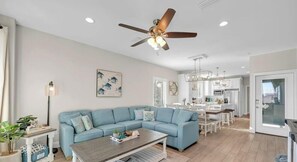 A living room with a blue sofa, a coffee table, a ceiling fan, and a kitchen in the background.