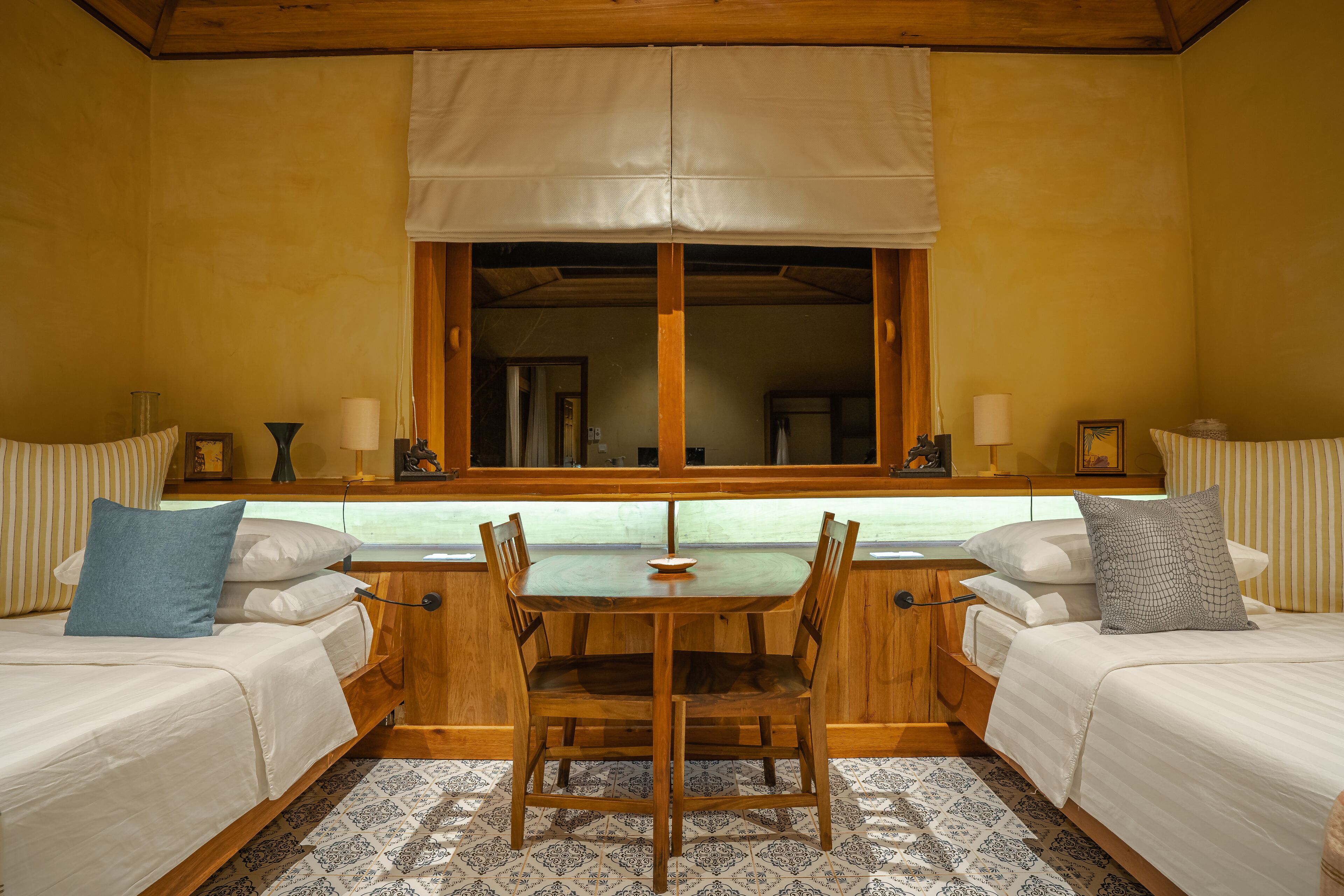 A room with two beds, a wooden table, and a window with a white valance.