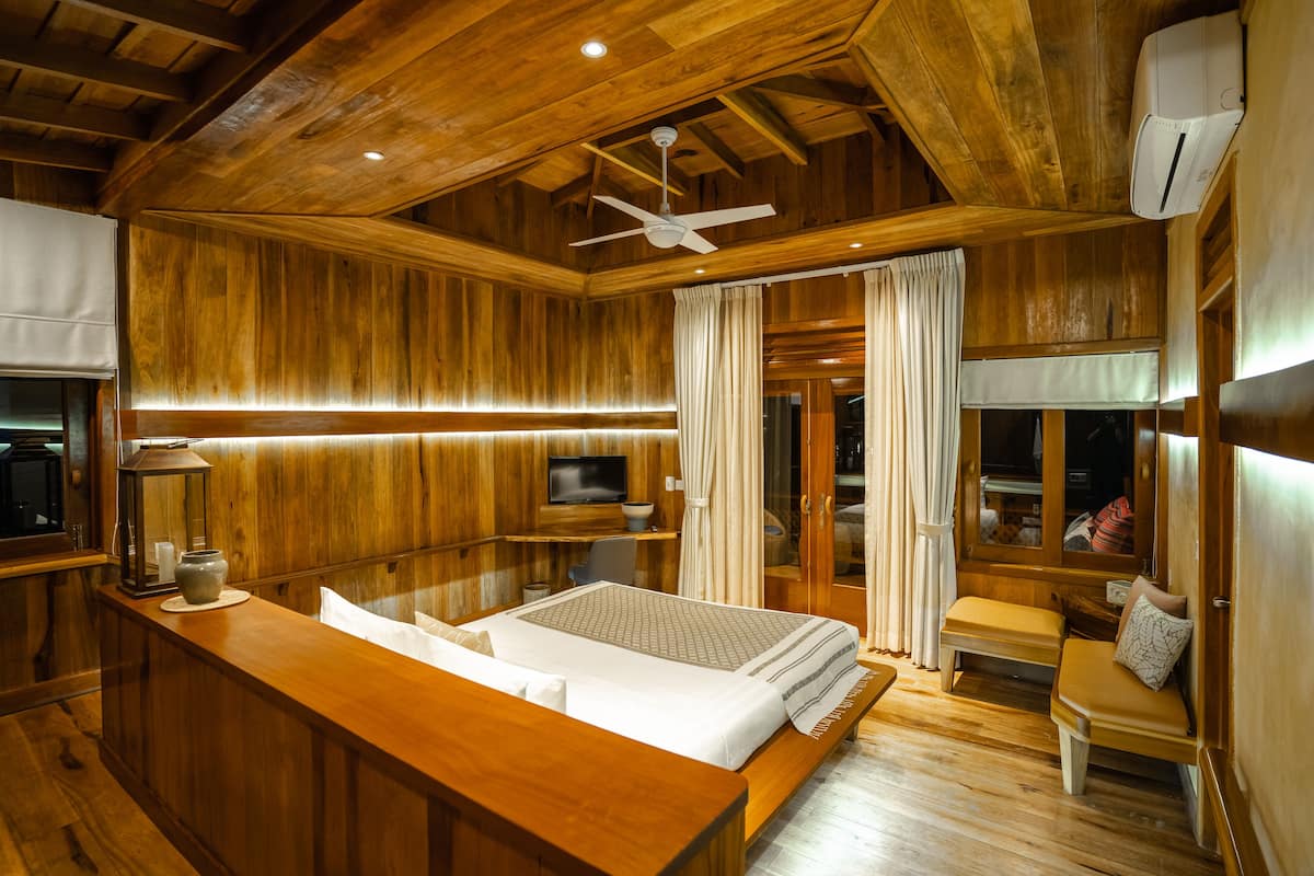 A wooden cabin interior with a bed, a desk, a television, and a ceiling fan.