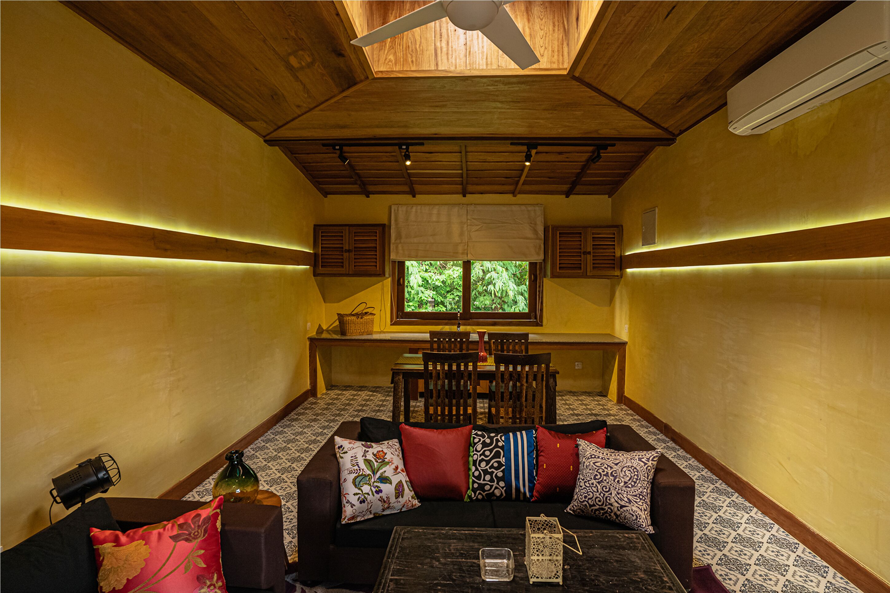 A cozy room with a wooden ceiling, a ceiling fan, a window with a view of greenery, a sofa with patterned cushions, and a small table with a candle and a glass.