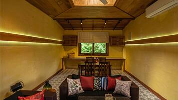 A cozy room with a wooden ceiling, a ceiling fan, a window with a view of greenery, a sofa with patterned cushions, and a small table with a candle and a glass.