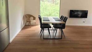A modern dining area with a wooden table and chairs, a wicker chair, a TV mounted on the wall, and a large window with a view of greenery. - Tenscott in Halls Gap (Halls Gap)