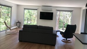 A modern living room with a large grey sofa, a black armchair, and a wooden rocking chair. There is a flat-screen TV mounted on the wall and a small table with a lamp. - Tenscott in Halls Gap (Halls Gap)