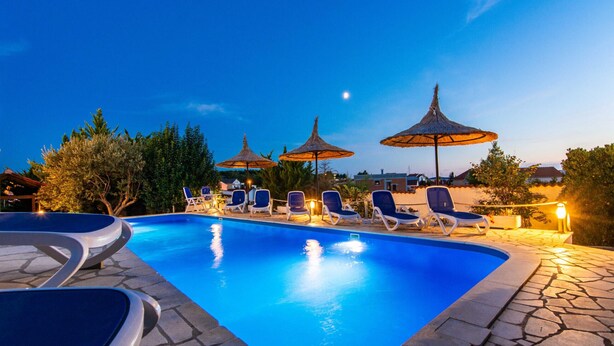 A swimming pool with lounge chairs and umbrellas, surrounded by trees and a clear night sky.