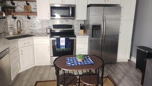 A modern kitchen with white cabinets, a stainless steel refrigerator, a microwave, and an oven. There is a small table with a placemat and a potted plant.