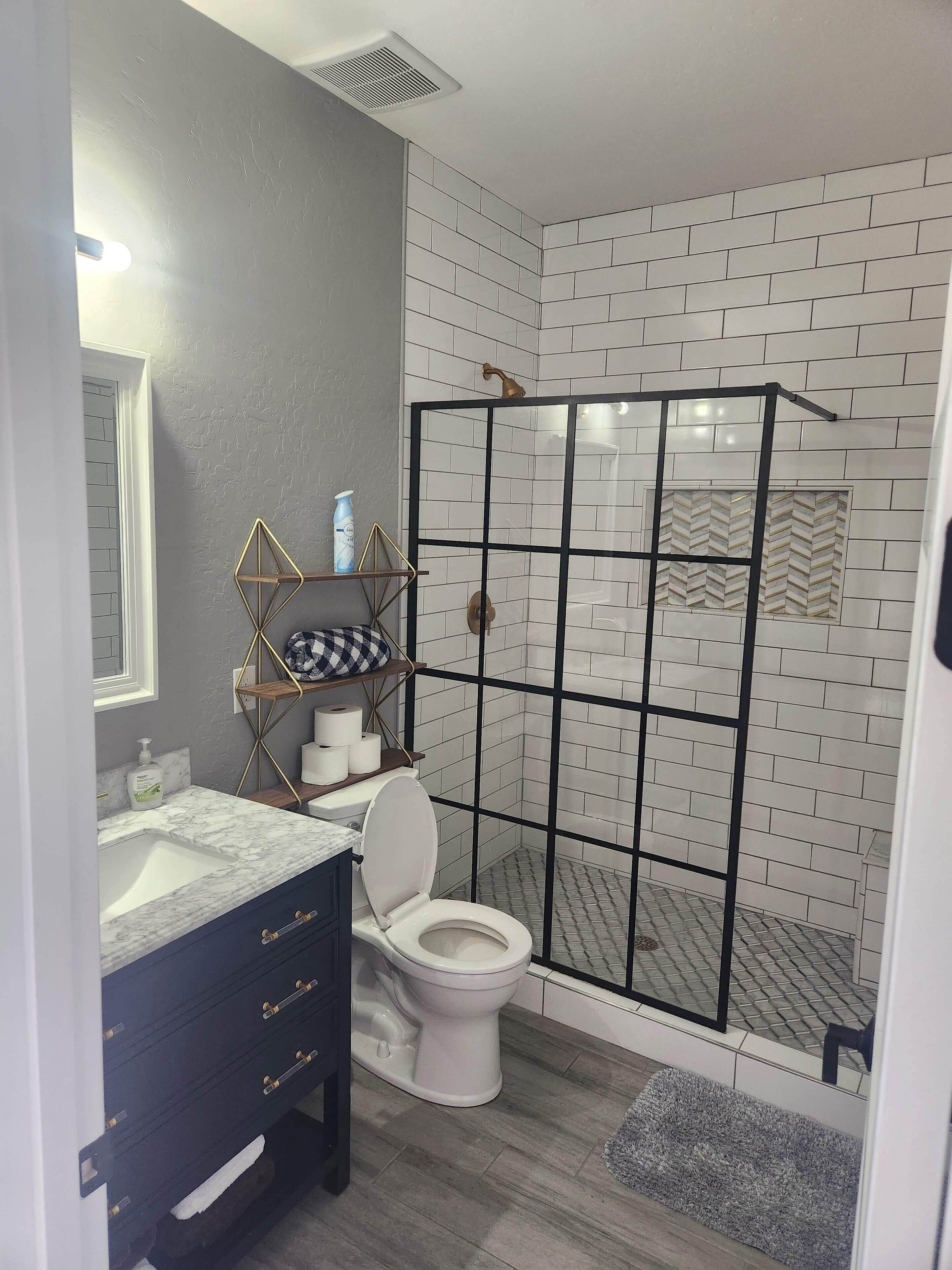 A bathroom with a white toilet, a marble sink, a dark wooden cabinet, a shower with a black metal frame, and a tiled wall with a herringbone pattern.