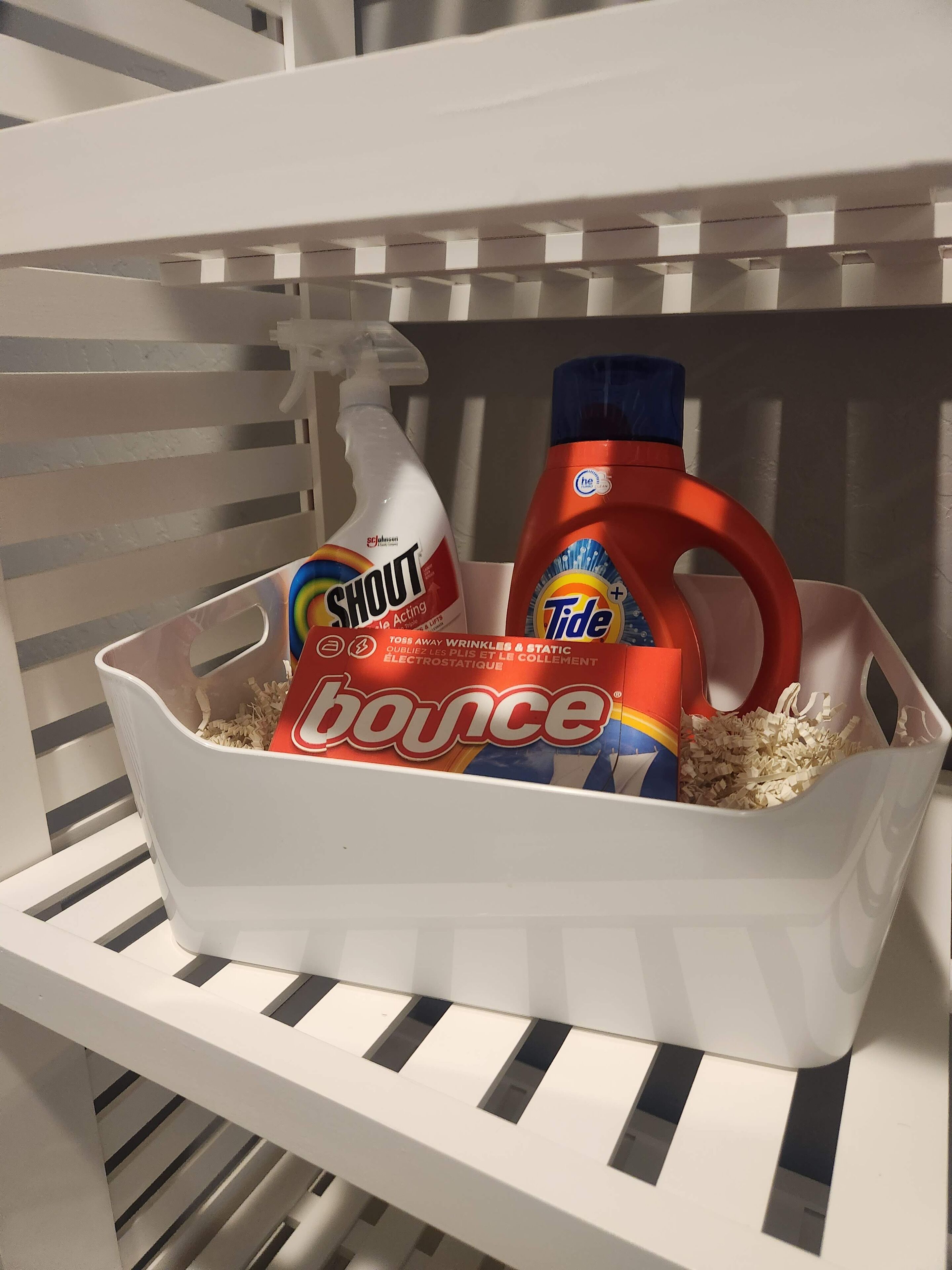 Un panier en plastique blanc contenant un flacon de liquide vaisselle Shout, un flacon de lessive Tide et un bidon de produit nettoyant pour sols Bounce.