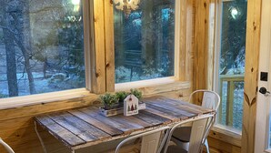 Un interior de madera con una mesa y sillas rústicas, un pequeño adorno con forma de casa, y vistas a un paisaje nevado a través de amplias ventanas.