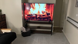 Una sala de estar con televisor mostrando una chimenea, un sofá oscuro, un pequeño otoman y un radiador blanco.