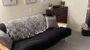 Una sala moderna con un sofá negro, una manta tejida y una pequeña mesa de madera. Hay dos impresiones botánicas enmarcadas en la pared, un mueble de madera oscura y una planta en maceta sobre una mesita.