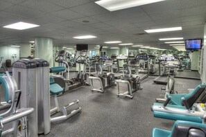 Un gimnasio con diversas máquinas de ejercicio, espejos y una pantalla de televisión.