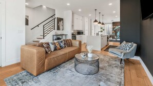A modern living room with a sofa, armchair, and coffee table. A staircase leads to a kitchen and dining area. - Group-Friendly 2 Nashville Homes • Sleeps 15 (Nashville)