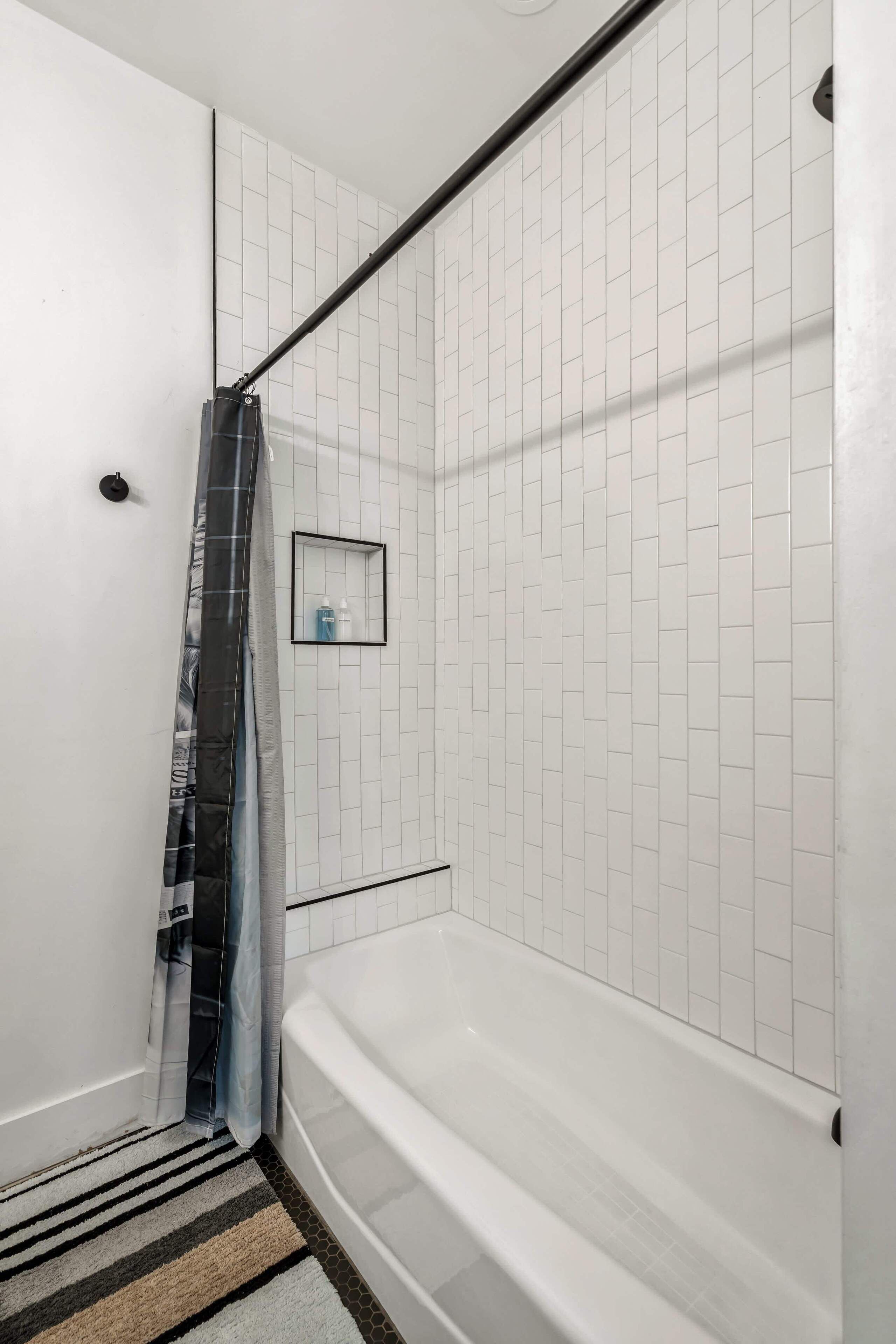 A bathroom with white tiled walls, a bathtub, a striped rug, and a shower curtain.