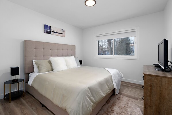 A bedroom with a large bed, a nightstand with a lamp, a dresser, a television, and a window with a view of trees.
