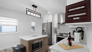 Et moderne køkken med indbygget ovn, mikroovn og rustfrit stål køleskab. Der er en træø med skærebræt og glas, samt en velkomstplakat på væggen.