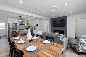 A modern kitchen with a built-in oven, a dining area with a wooden table set for four, and a grey sofa in the living area.