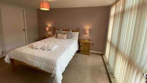 A neatly made bed with a white comforter and pillows, a bedside table with a lamp, and a window with sheer curtains.